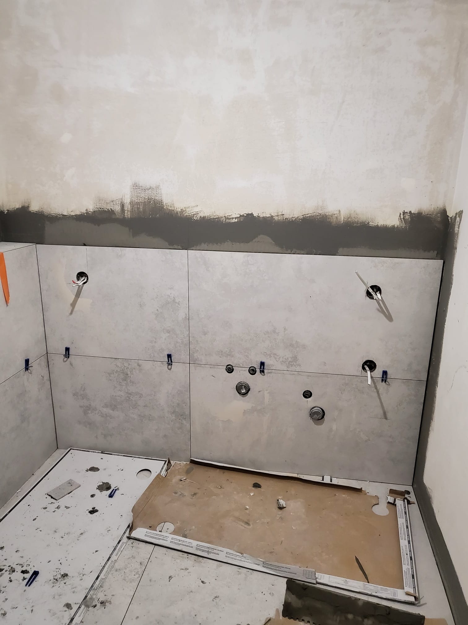 Ściana łazienki w trakcie układania jasnych płytek. Widoczne otwory na instalacje, poziomica laserowa i plastikowe krzyżyki dystansowe.