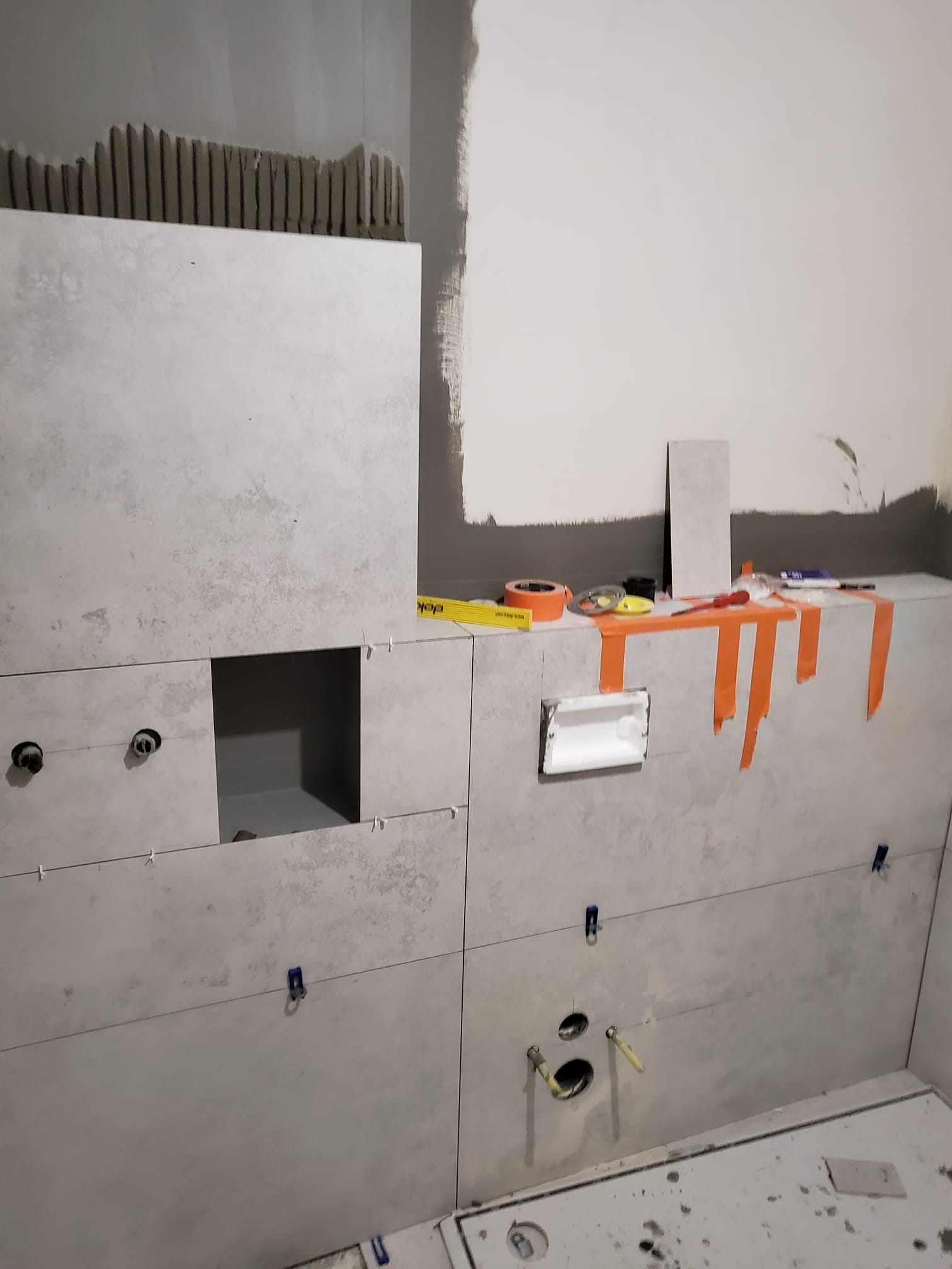 Montaż płytek w łazience, widoczne narzędzia i akcesoria glazurnicze, otwory na armaturę, jasne płytki z delikatnym wzorem. Etap prac wykończeniowych.