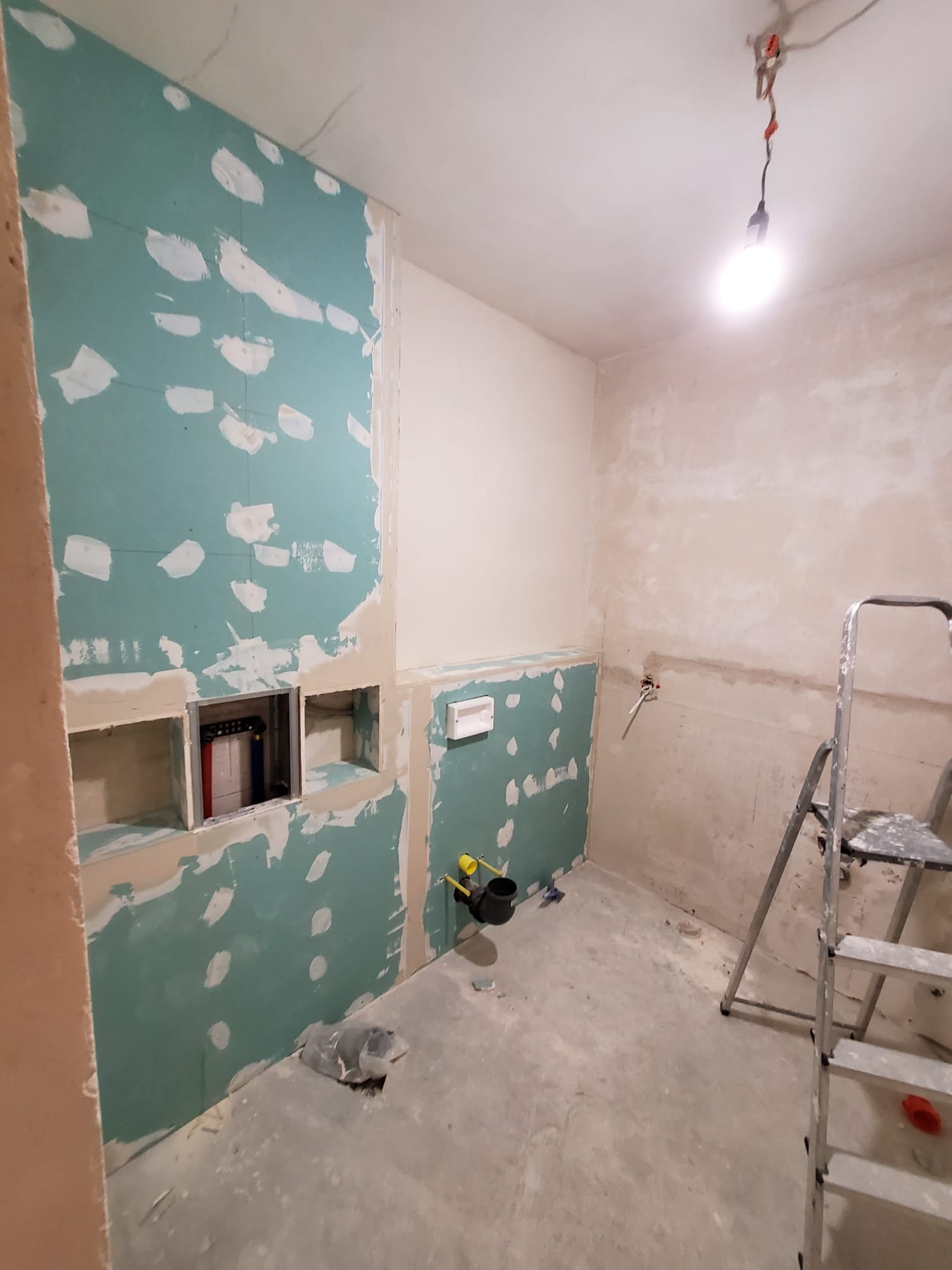 Łazienka w trakcie prac: płyty gips-karton na ścianach, widoczne instalacje, drabina. W ścianie wnęki na półki. Surowy stan, przygotowanie do dalszych prac wykończeniowych.