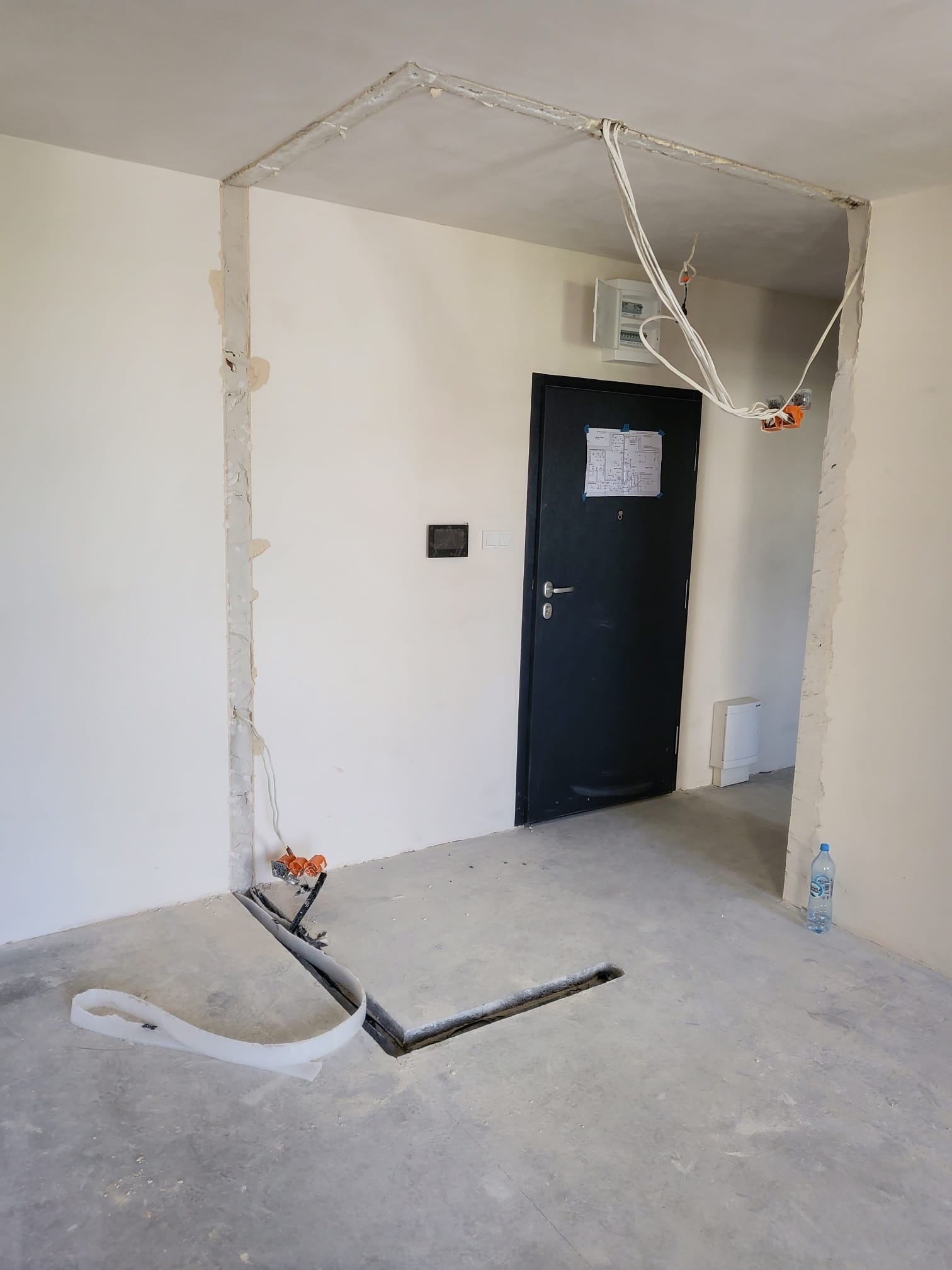 Wnętrze w trakcie remontu: widoczne kable, bruzdy w ścianach i podłodze, instalacja elektryczna, drzwi wejściowe. Butelka wody przy ścianie.