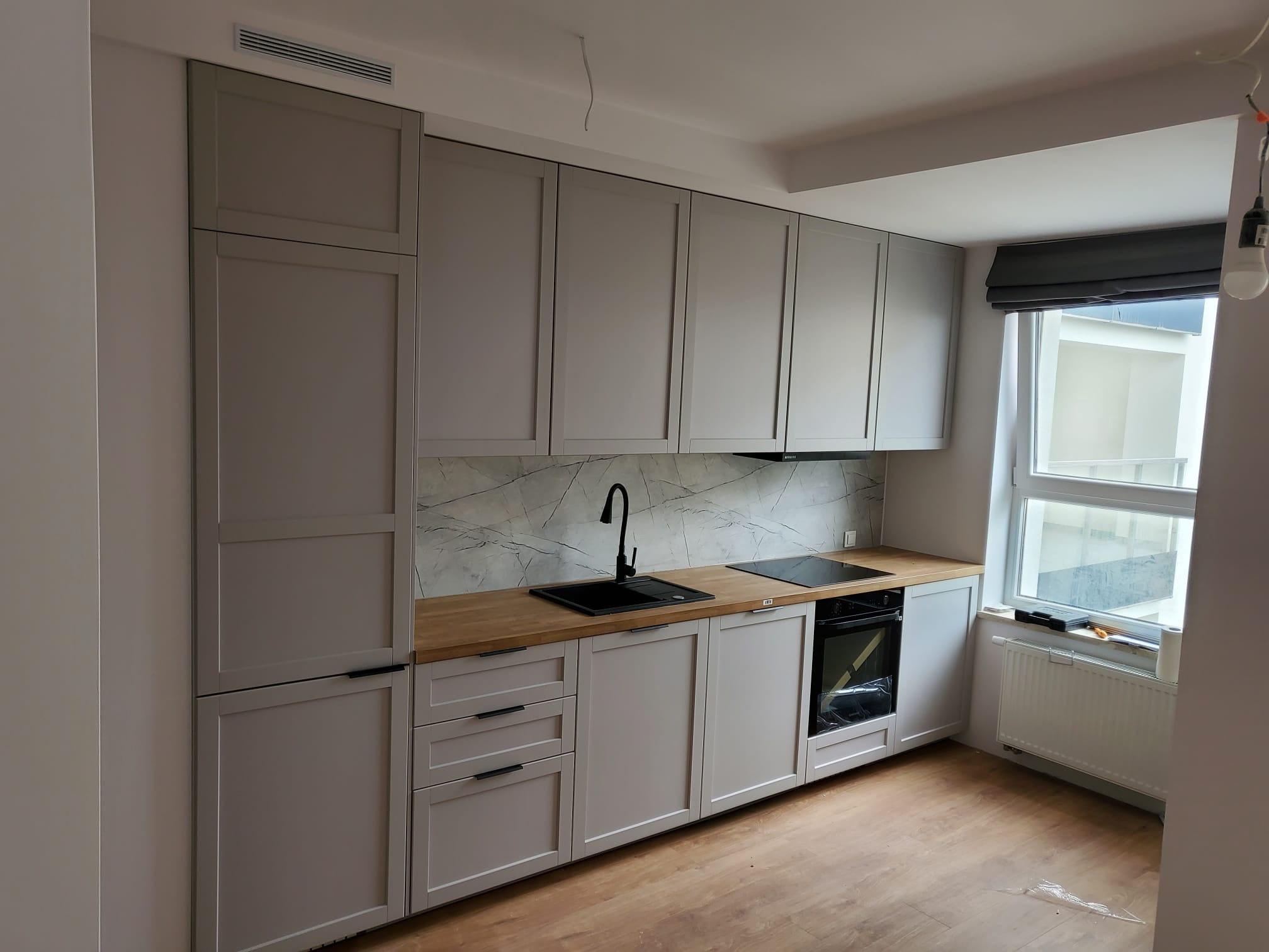 Jasna kuchnia z szarymi szafkami, drewnianym blatem i marmurowym backsplashiem. Czarny zlew i płyta grzewcza. Okno z roletą rzymską. Nowoczesny design.