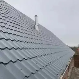 Szary dach z blachy trapezowej z kominem, widok z dołu, na tle pochmurnego nieba. Widoczny fragment muru budynku.