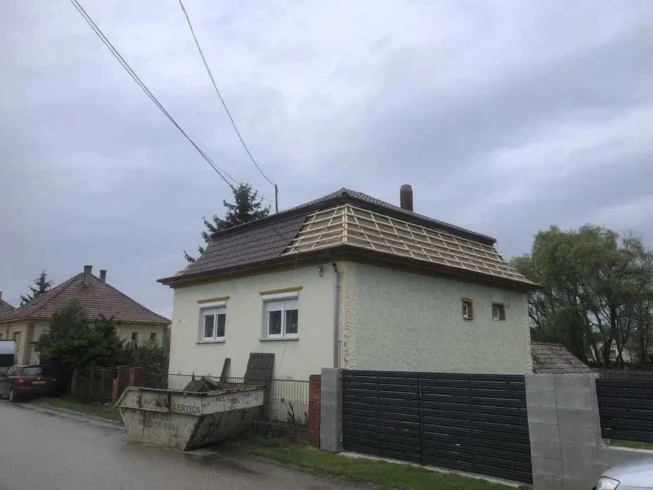 Dom jednorodzinny z częściowo zdjętym dachem, widoczna więźba. Kontener na odpady budowlane przed budynkiem. Pochmurne niebo w tle.