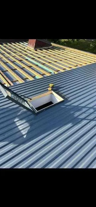 Montaż okna dachowego na blaszanym dachu w trakcie budowy. Widoczne drewniane łaty i fragment komina w tle. Użyto wkrętarki.