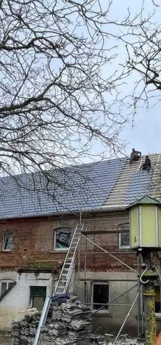 Prace dekarskie na dachu starego domu z cegły. Na dachu dwaj dekarze układają dachówkę. Na dole rusztowanie, drabina i stos dachówek.