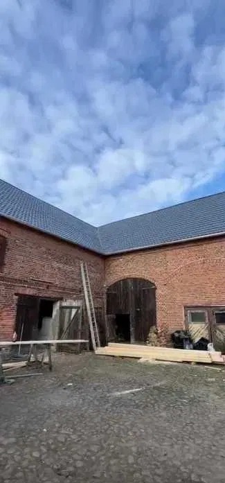 Ceglany budynek gospodarczy z nowym, ciemnym dachem pod błękitnym niebem. Widoczna drabina i materiały budowlane. Kąt ujęcia z dołu podkreśla wysokość budynku.