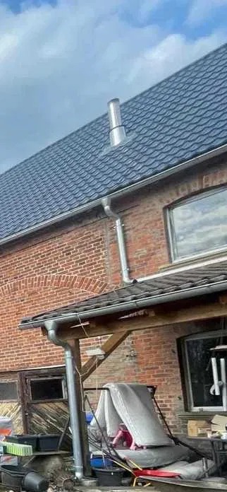 Dom z ciemną dachówką i metalowym kominem, widoczne rynny i ceglana ściana. Część dachu zadaszona. Ujęcie z dołu, widoczne niebo.