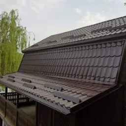 Ciemnobrązowy dach z blachy trapezowej na drewnianej altanie, widok z dołu, z rynną i elementami wentylacyjnymi. W tle drzewo i pochmurne niebo.