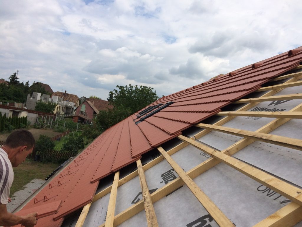 Częściowo pokryty dach czerwoną dachówką z widoczną drewnianą konstrukcją więźby i oknem dachowym na tle zabudowań i pochmurnego nieba. Prace dekarskie w toku.