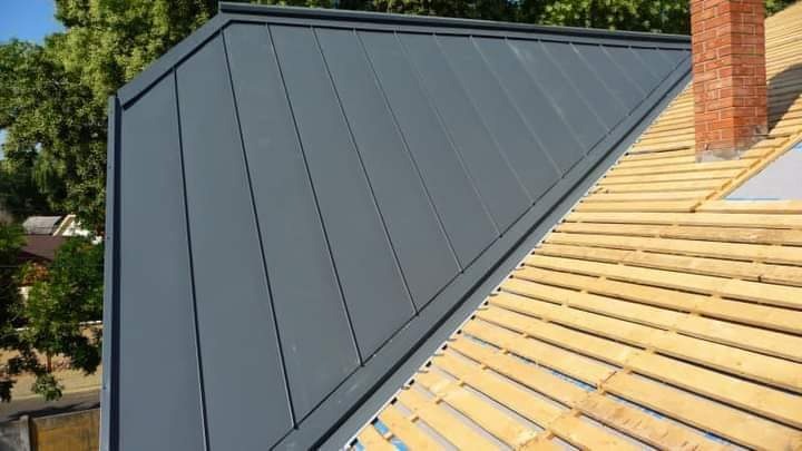 Częściowo pokryty dach blachą w kolorze grafitowym, z widocznym kominem z cegły i drewnianą konstrukcją pod pokrycie. Widok z góry, słoneczny dzień.