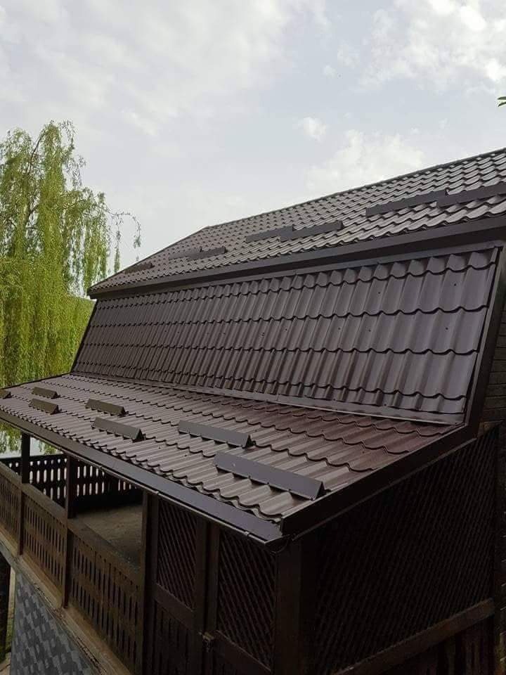 Ciemnobrązowy dach z blachy trapezowej na drewnianej altanie, widok z dołu, z rynną i elementami wentylacyjnymi. W tle drzewo i pochmurne niebo.