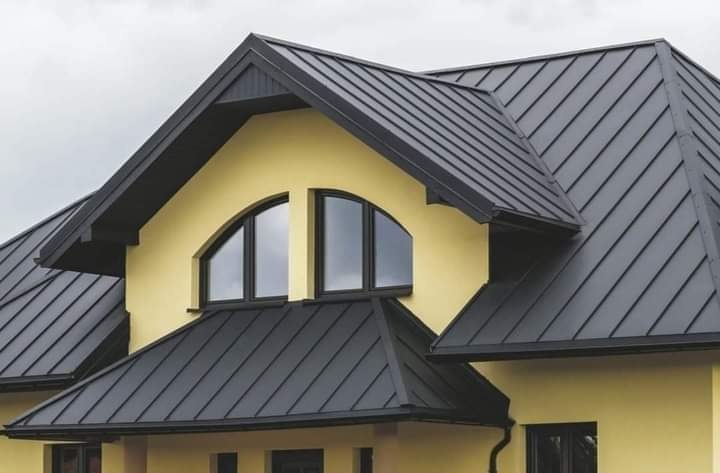 Żółty dom z czarnym dachem z blachy na rąbek stojący. Widoczne dwa okna w kształcie łuku i rynny. Nowoczesne krycie dachu.