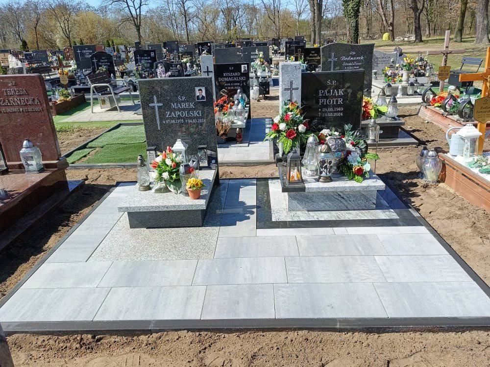 Dwa nagrobki z szarego granitu, otoczone jasnoszarą kostką brukową na cmentarzu. Kompozycje kwiatowe i znicze zdobią pomniki. Słoneczny dzień.
