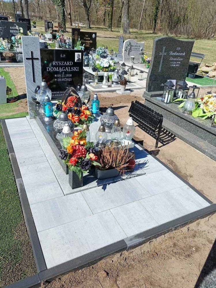 Granitowy nagrobek z szarych płyt brukowych, otoczony czarnym obramowaniem. Na nagrobku znicze, kwiaty i zdjęcie zmarłego. W tle inne groby i drzewa.