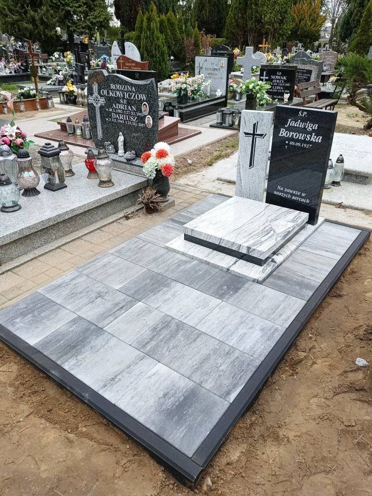 Nowo ułożona, szara kostka brukowa wokół nagrobka na cmentarzu. Prostokątne płyty ułożone równo, obramowane czarną obwódką. W tle inne nagrobki i zieleń.