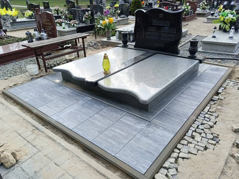 Granitowy nagrobek z szarą płytą i czarną tablicą, otoczony kostką brukową w trakcie układania, cmentarz w tle.
