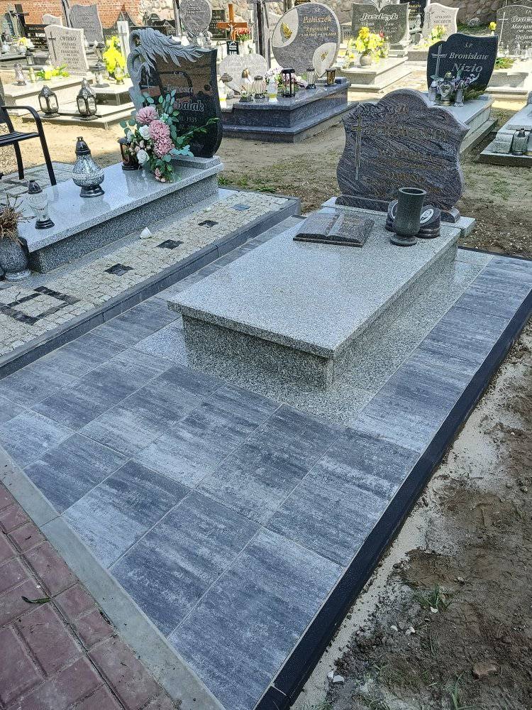 Granitowy nagrobek z książką i wazonem, otoczony szarą kostką brukową na cmentarzu. Widok z bliska na nowo ułożoną nawierzchnię.