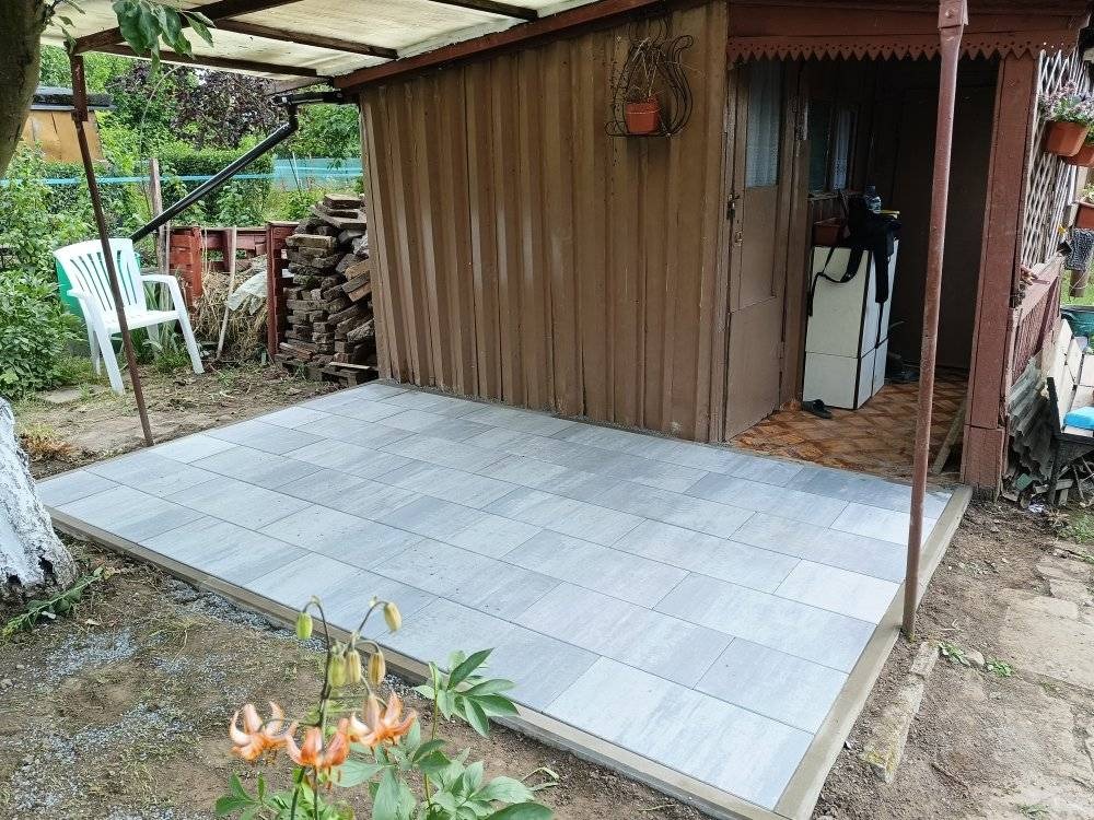 Nowy taras z jasnoszarej kostki brukowej przy drewnianym domku, z widocznymi kwiatami na pierwszym planie i stertą cegieł w tle, w naturalnym oświetleniu.