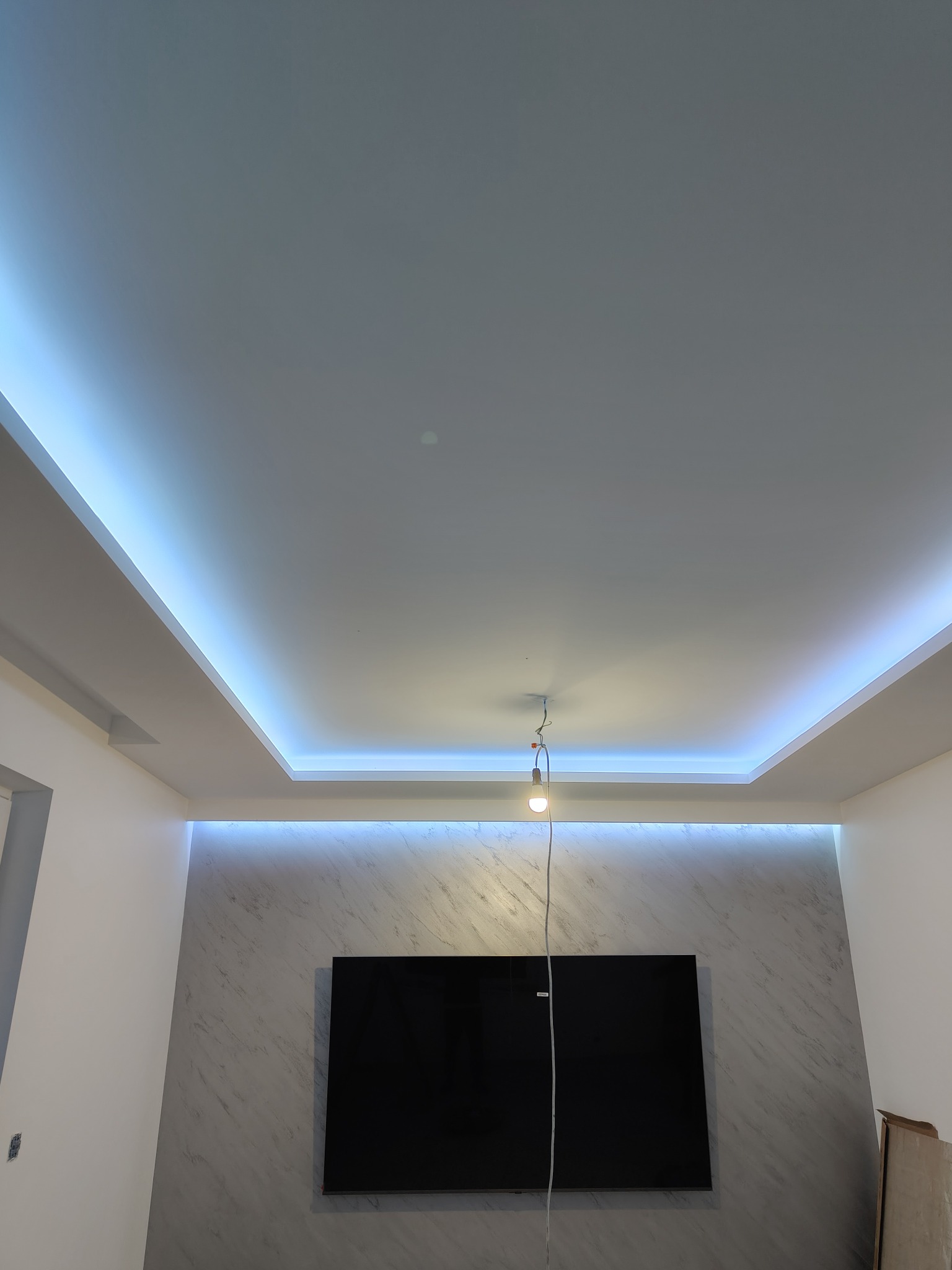 Sufit podwieszany z oświetleniem LED w nowoczesnym salonie. Dekoracyjna ściana z efektem marmuru, centralnie umieszczony telewizor. W tle widoczne elementy wykończenia.
