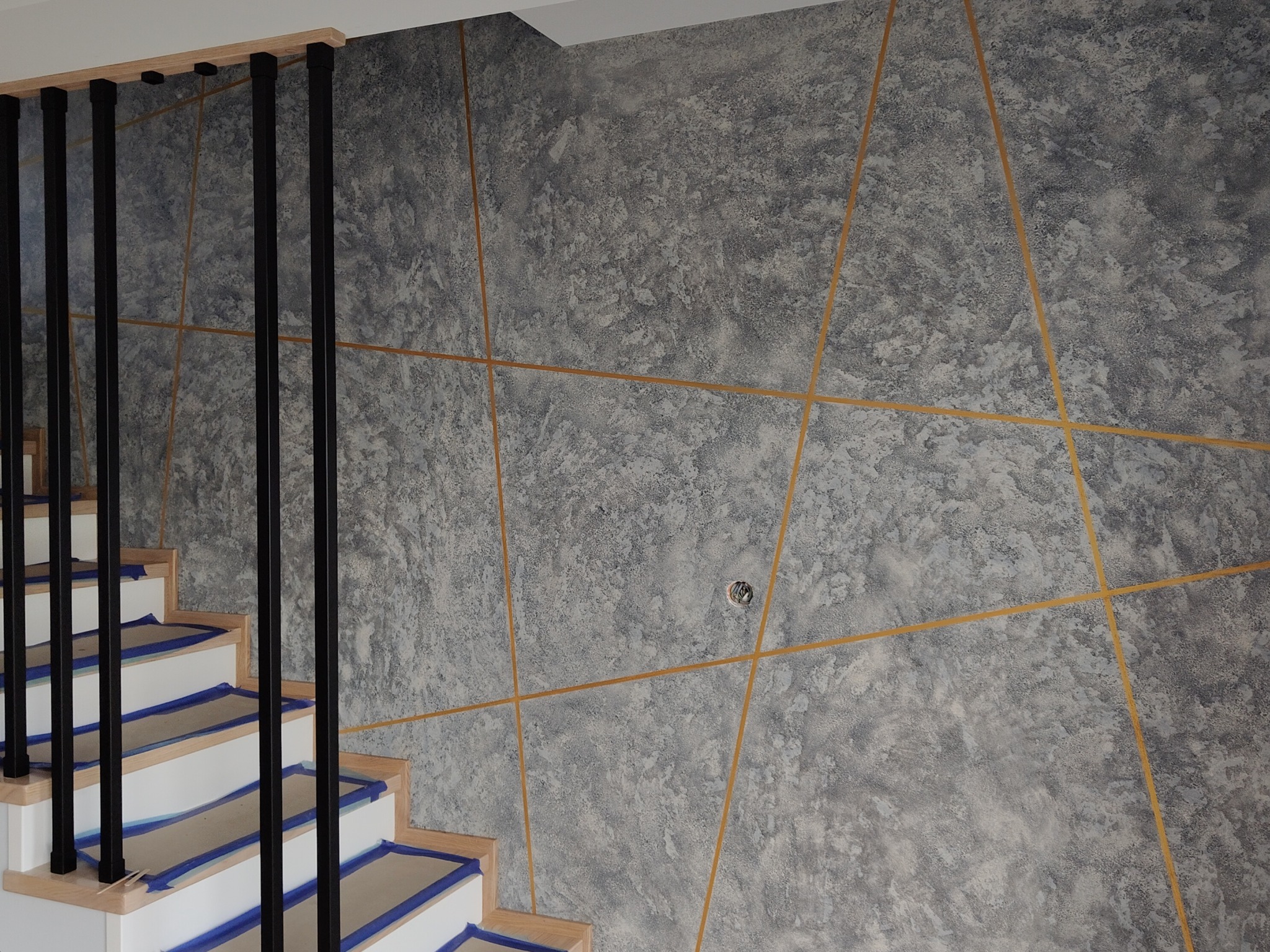 Dekoracyjna ściana w szarościach z geometrycznym wzorem, widok na fragment schodów z czarną balustradą. Unikalna faktura ściany i złote linie tworzą nowoczesny design.