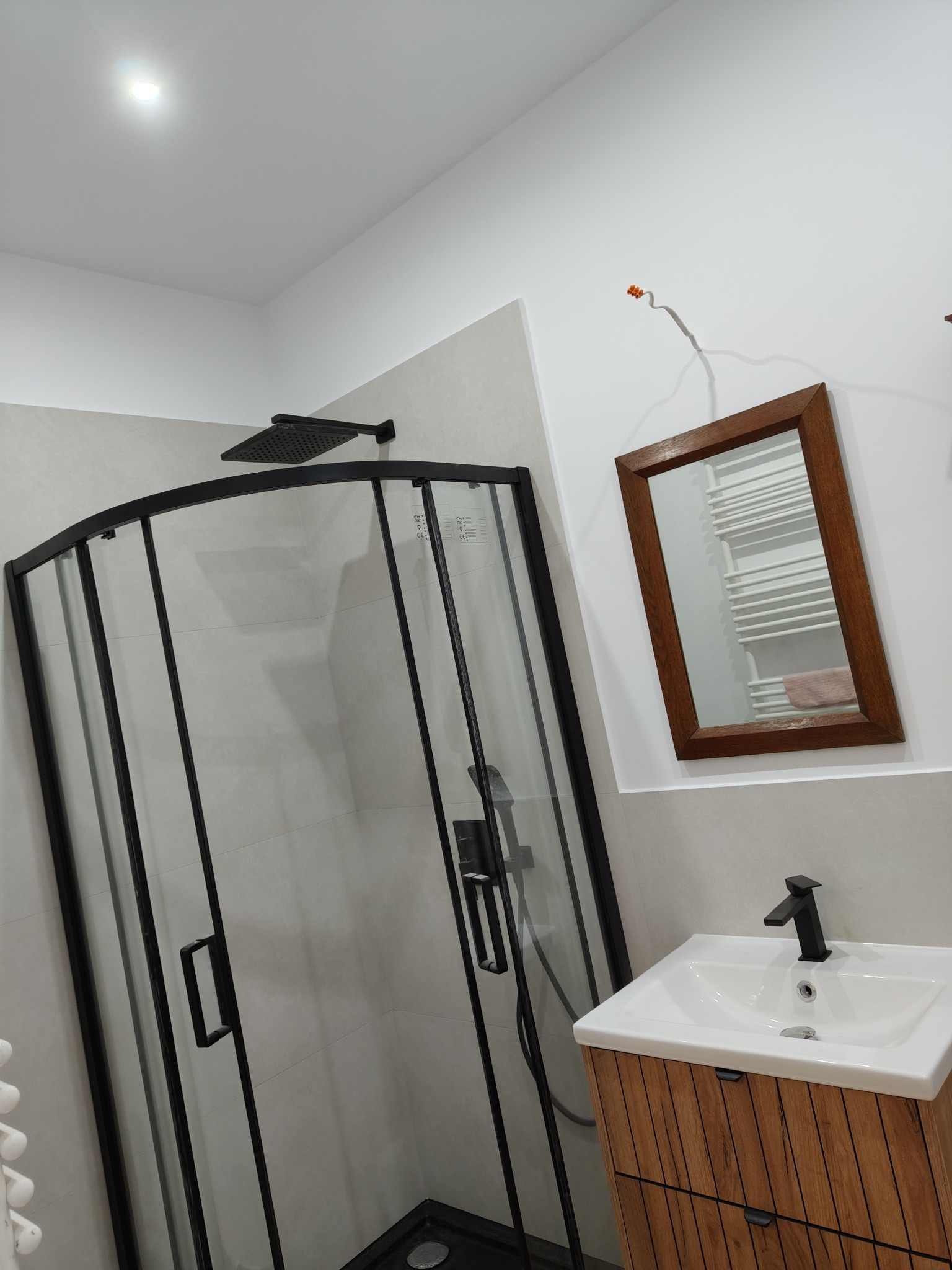 Nowoczesna łazienka z czarną kabiną prysznicową, umywalką na drewnianej szafce i lustrem w drewnianej ramie. Minimalistyczny wystrój z akcentem na funkcjonalność.