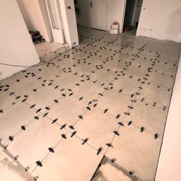 Reli.com.pl - Układanie beżowych płytek na podłodze z użyciem czarnych krzyżyków dystansowych, widoczne niedokończone fragmenty i białe ściany w tle.