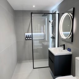 Reli.com.pl - Nowoczesna łazienka w szarościach z czarnymi akcentami: kabina prysznicowa typu walk-in, podświetlane lustro i umywalka z szafką.