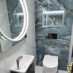 Reli.com.pl - Nowoczesna łazienka z umywalką na szafce, podświetlanym lustrem i płytkami imitującymi marmur. W tle toaleta i dekoracyjna wnęka z oświetleniem.