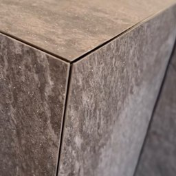 Reli.com.pl - Szczegółowe ujęcie narożnika blatu wykończonego szarymi płytkami imitującymi kamień, z widocznymi fugami i delikatnym połyskiem.