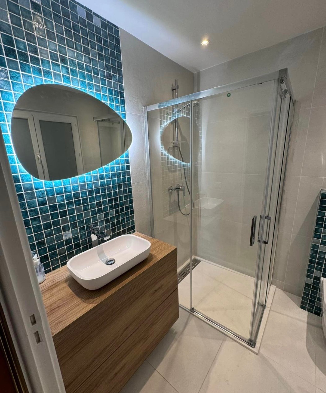 Nowoczesna łazienka z kabiną prysznicową i umywalką nablatową na drewnianej szafce. Ściana w turkusowej mozaice z podświetlanym owalnym lustrem.