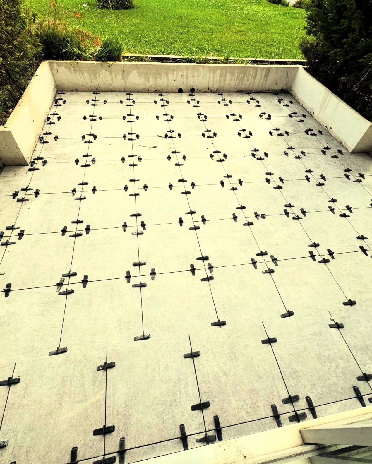 Przygotowanie do układania płytek na tarasie z użyciem systemu poziomowania. Widoczne krzyżyki dystansowe i kliny. Tło stanowi trawnik i zieleń.