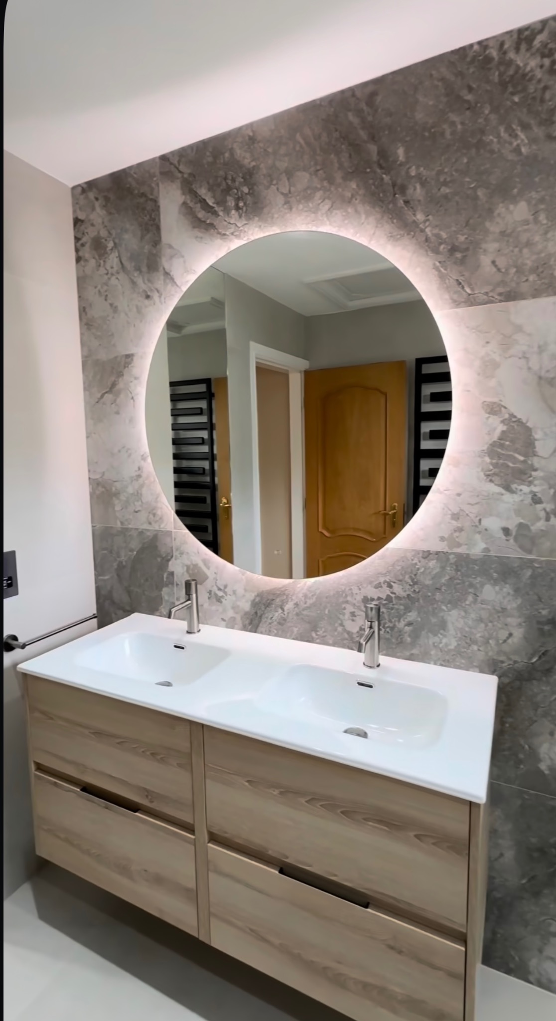 Stylowa łazienka we Wrocławiu: podwójna umywalka z szafką z jasnego drewna, okrągłe lustro z podświetleniem i marmurowe płytki ścienne.