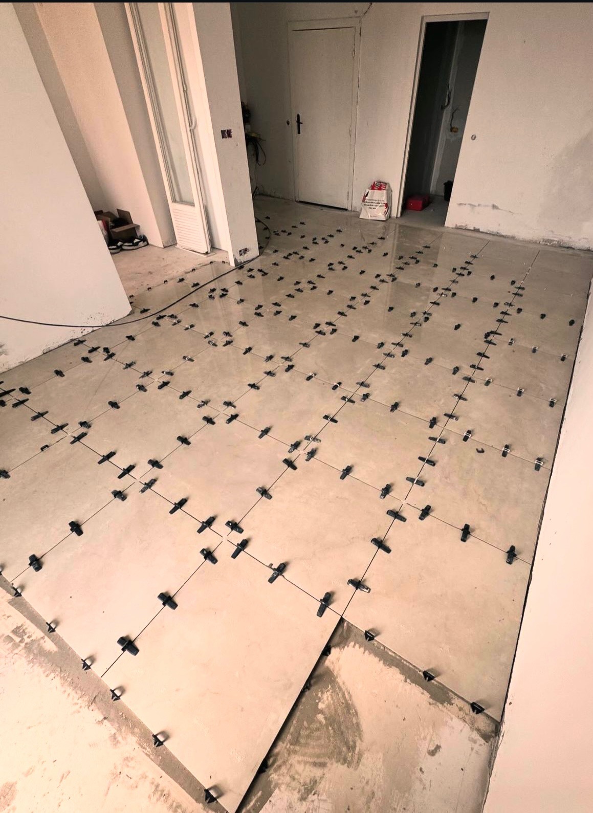 Układanie beżowych płytek na podłodze z użyciem czarnych krzyżyków dystansowych, widoczne niedokończone fragmenty i białe ściany w tle.