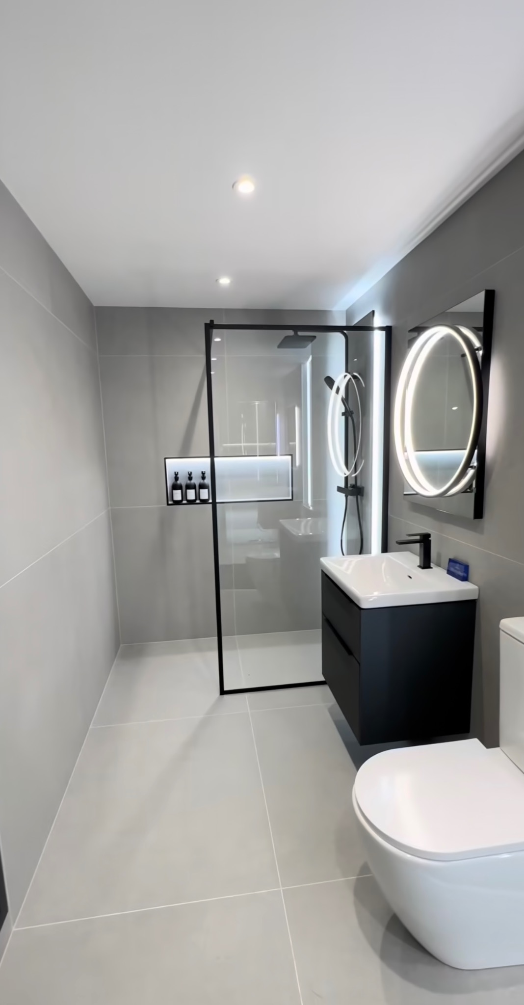 Nowoczesna łazienka w szarościach z czarnymi akcentami: kabina prysznicowa typu walk-in, podświetlane lustro i umywalka z szafką.