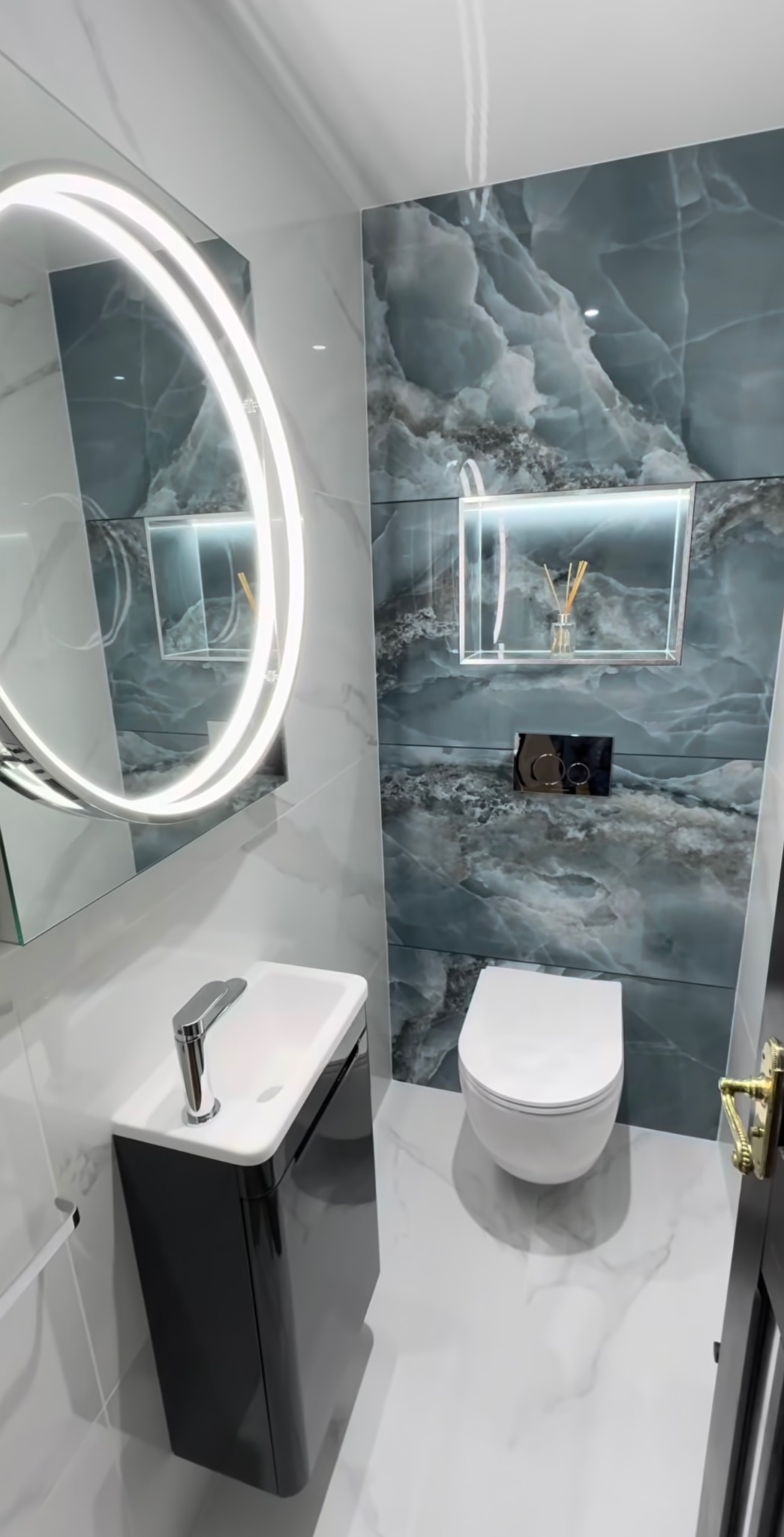 Nowoczesna łazienka z umywalką na szafce, podświetlanym lustrem i płytkami imitującymi marmur. W tle toaleta i dekoracyjna wnęka z oświetleniem.