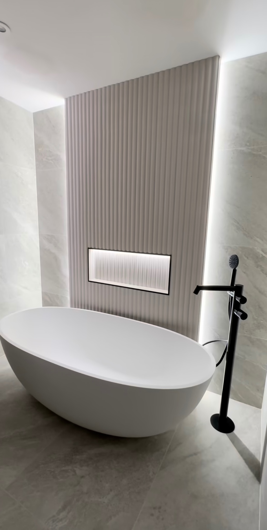 Minimalistyczna łazienka z wolnostojącą wanną i dekoracyjną ścianą z oświetleniem LED. Czarna bateria podłogowa dodaje elegancji wnętrzu.