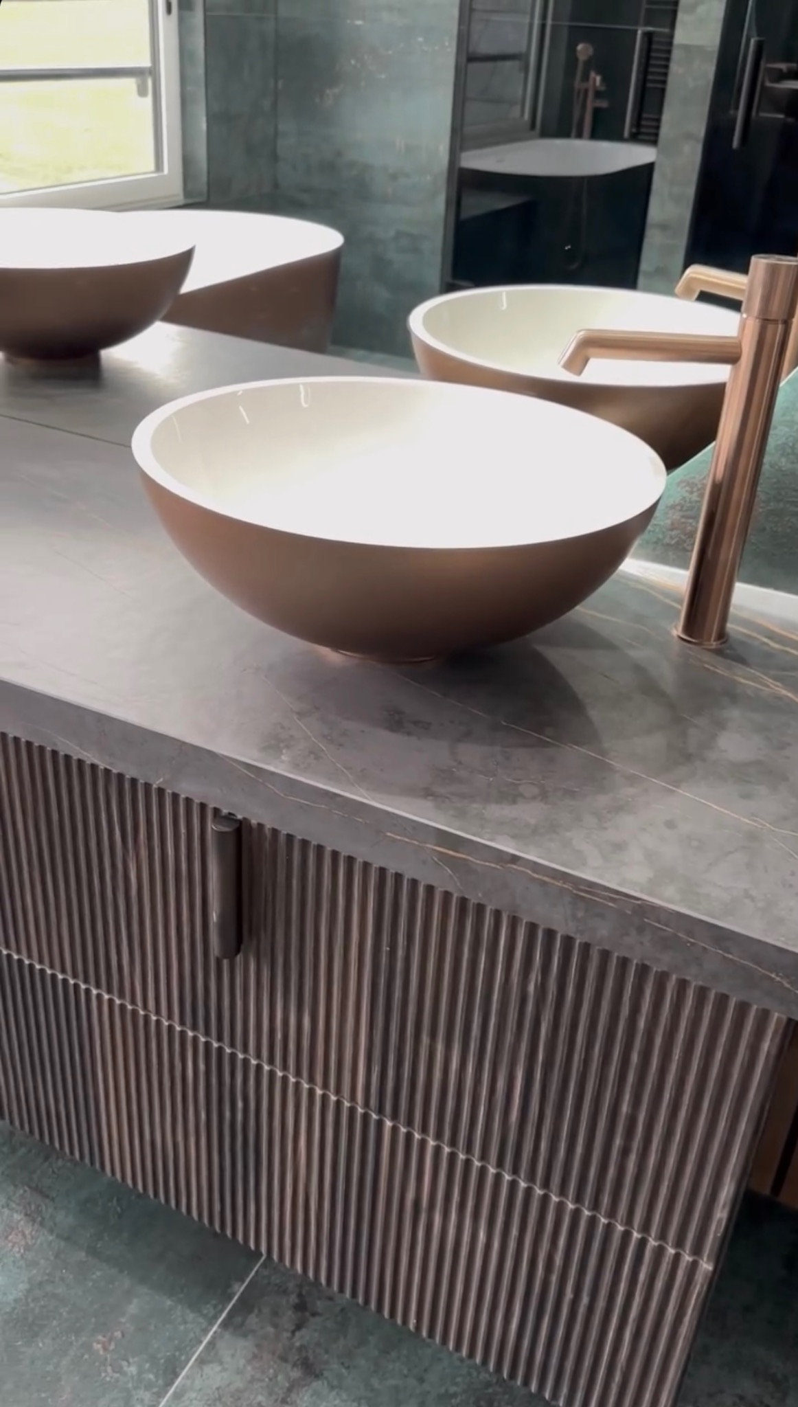 Nowoczesna łazienka z umywalkami w kształcie misy i armaturą w odcieniach miedzi. Blat z ciemnego kamienia z wyraźnym żyłowaniem. Szafka z lamelami.