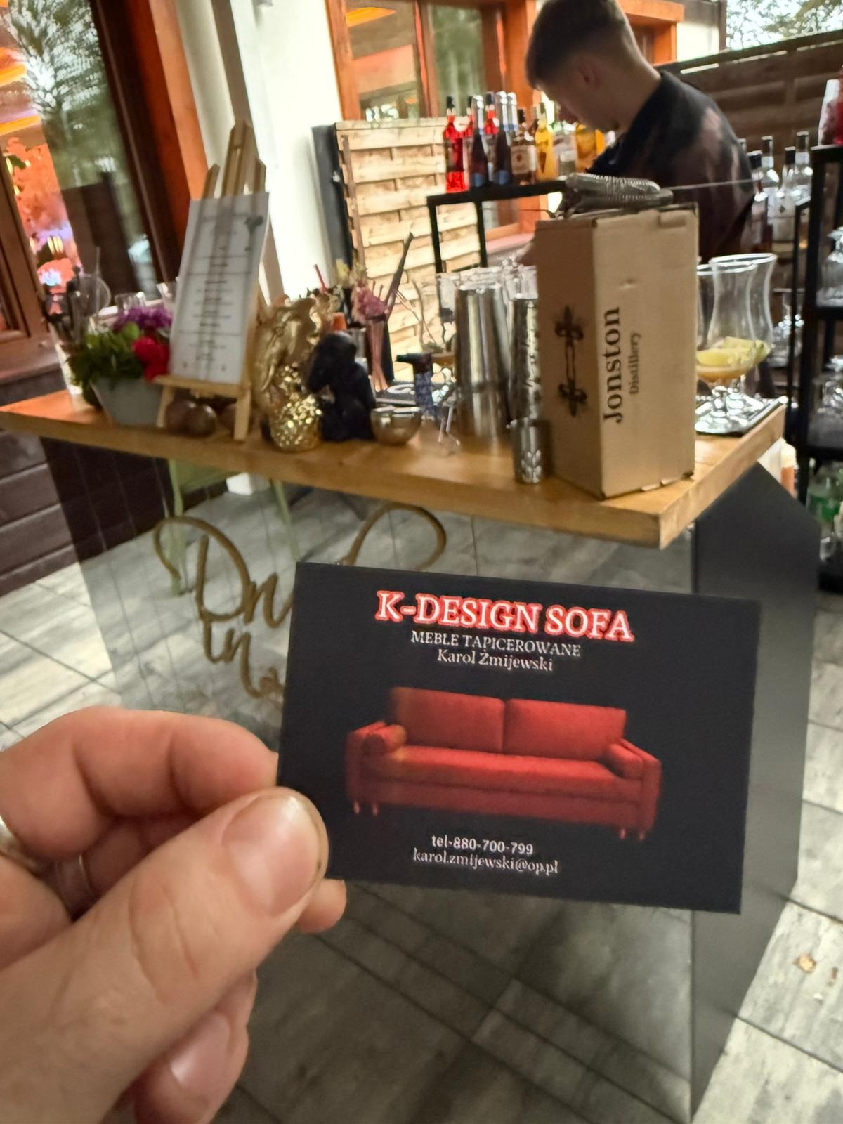 Wizytówka K-Design Sofa z ilustracją czerwonej sofy na tle baru z akcesoriami, prezentowana w dłoni. Meble tapicerowane Karol Zmijewski.