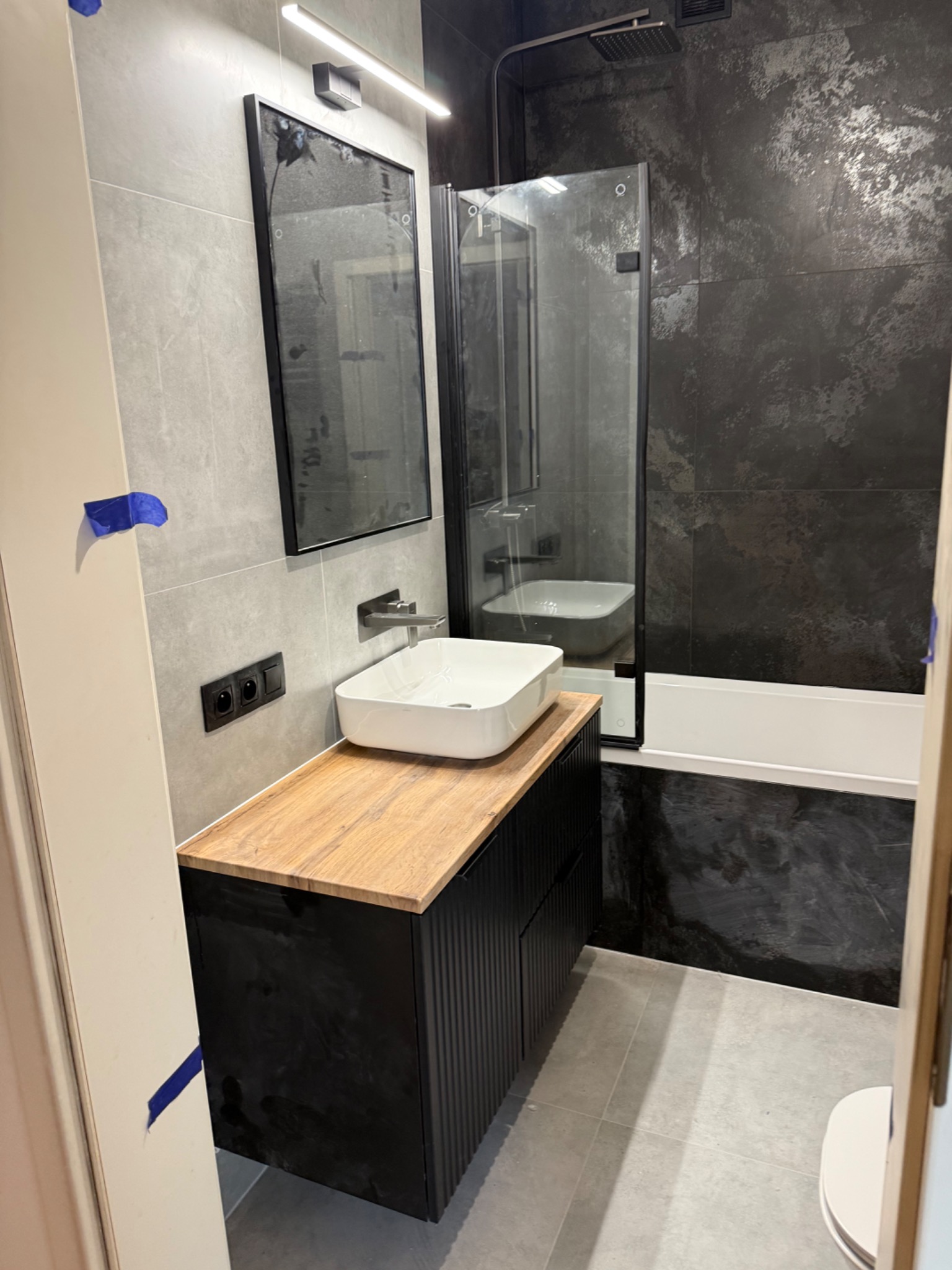 Nowoczesna łazienka: szare płytki, czarna armatura, drewniany blat pod umywalką i kabina prysznicowa z czarnymi profilami. Minimalistyczny design.