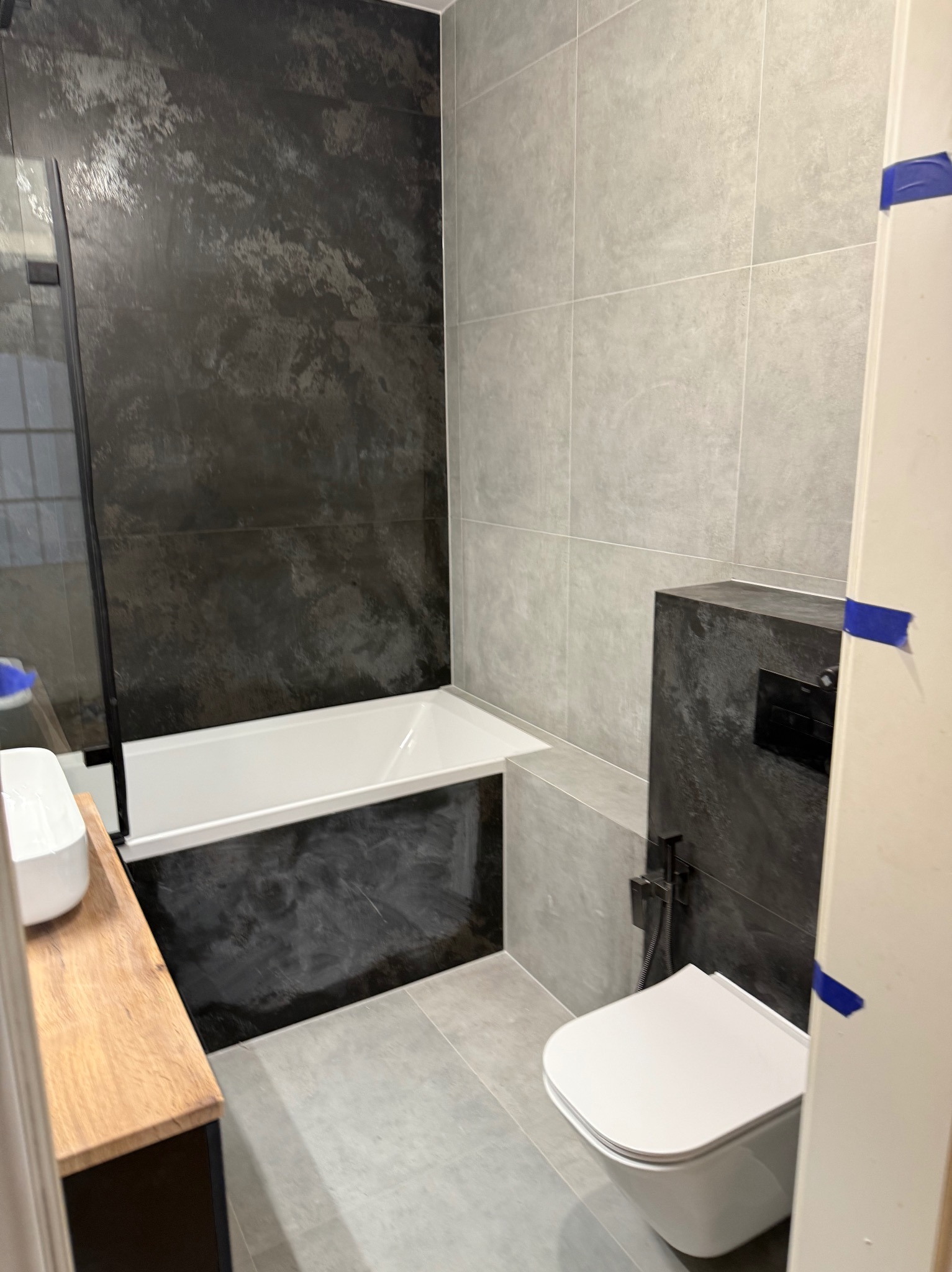 Nowoczesna łazienka po remoncie: czarno-białe płytki, wanna z parawanem, podwieszana toaleta, drewniany blat umywalki i szara podłoga.