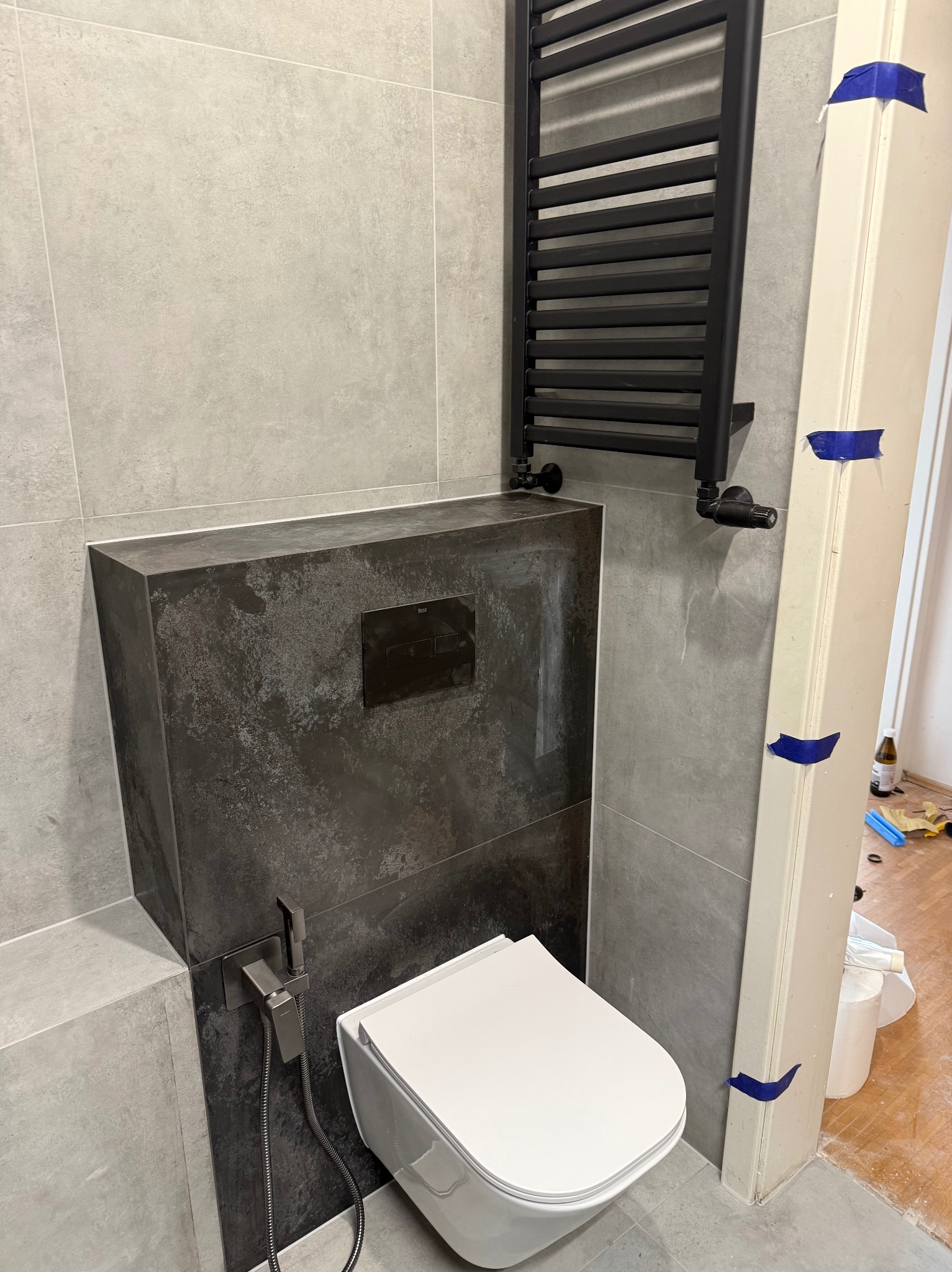 Nowoczesna łazienka w szarościach: podwieszana toaleta z bidetką, czarny grzejnik drabinkowy, płytki imitujące beton. Wykończenie z dbałością o detale.