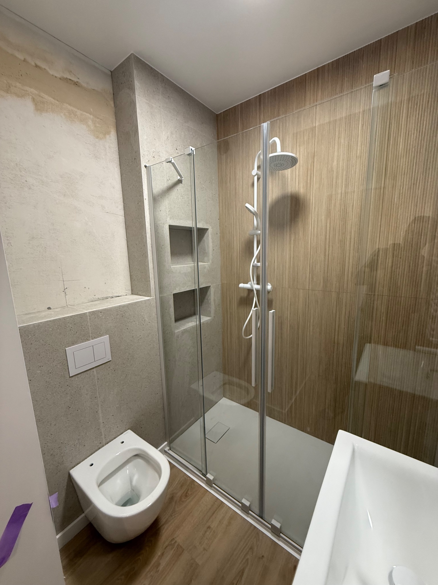 Nowoczesna łazienka w trakcie prac wykończeniowych: kabina prysznicowa ze szklanymi drzwiami, podwieszana toaleta i fragment ściany przed położeniem gładzi.