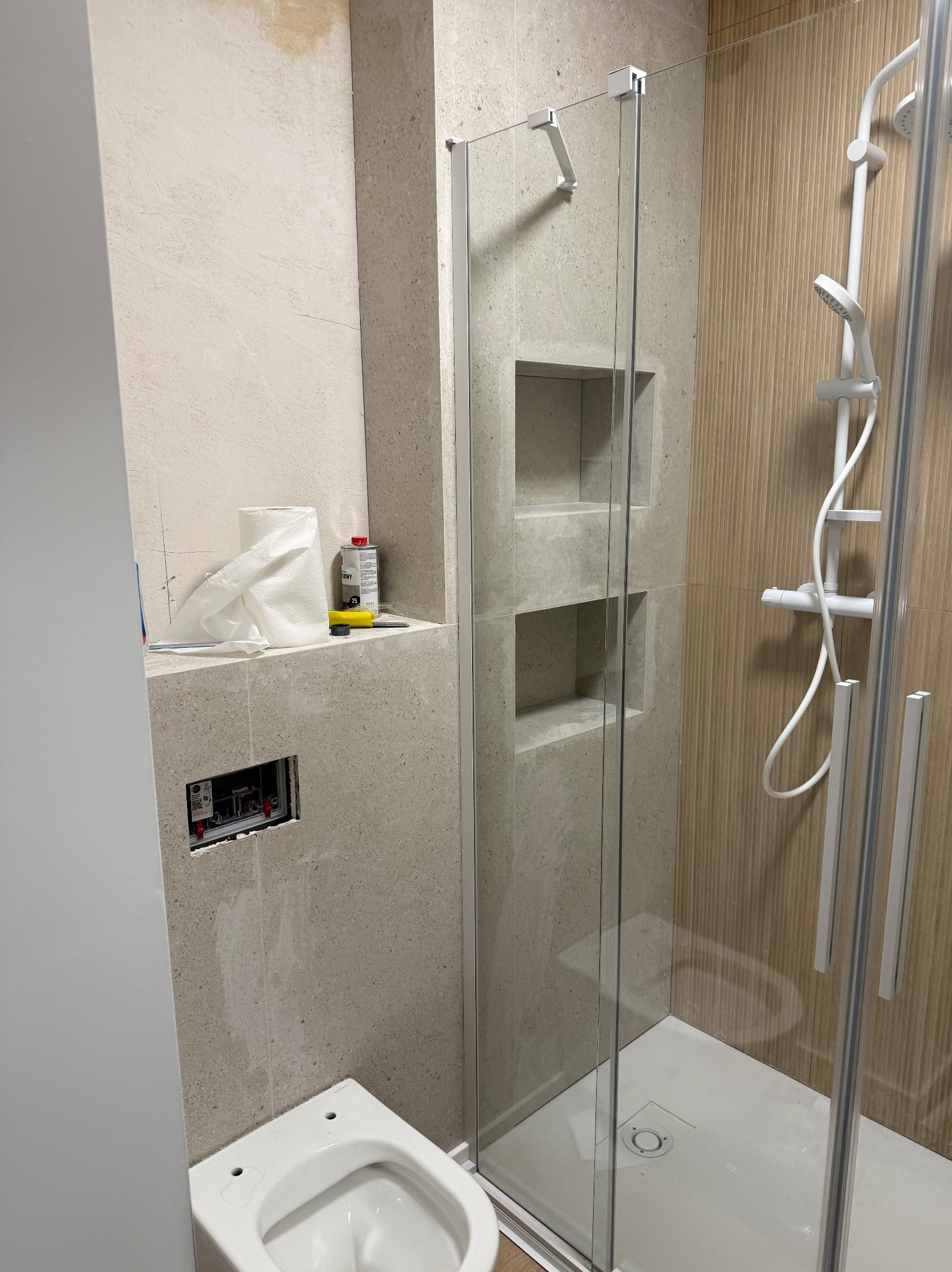 Nowa łazienka w trakcie wykańczania: kabina prysznicowa ze szklaną ścianką, częściowo zamontowana toaleta, widoczne narzędzia i materiały budowlane na półce.