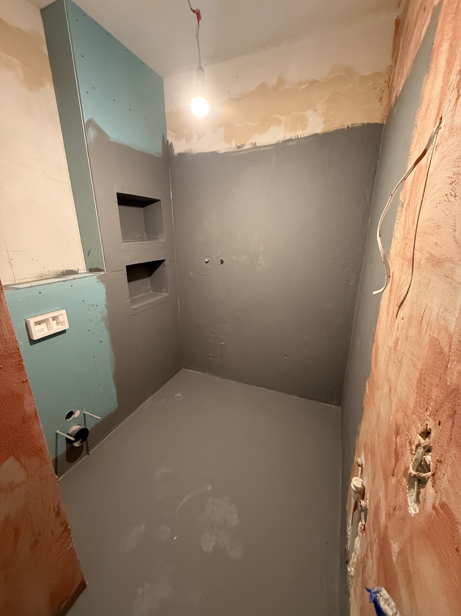 Łazienka w trakcie hydroizolacji: szare ściany i podłoga, wnęki w ścianie, niebieskie płyty gipsowe, widoczne instalacje. Etap przygotowania do dalszych prac.