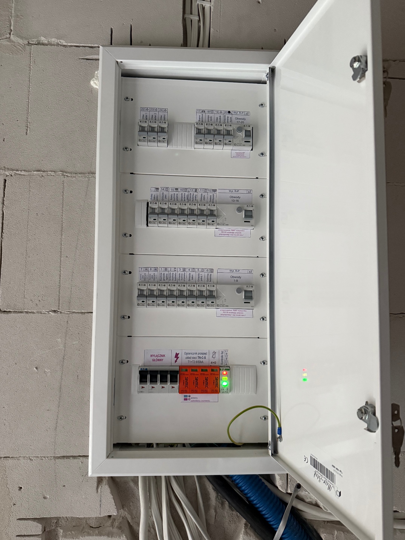 Otwarta skrzynka elektryczna z bezpiecznikami i wyłącznikami, widoczna na ścianie z betonu komórkowego. Widoczne przewody.
