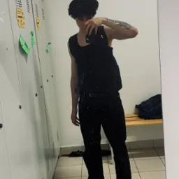 Mężczyzna w czarnym ubraniu robi selfie w lustrze na tle szatni z metalowymi szafkami i ławką. Widoczne tatuaże na ramionach i białe buty sportowe.