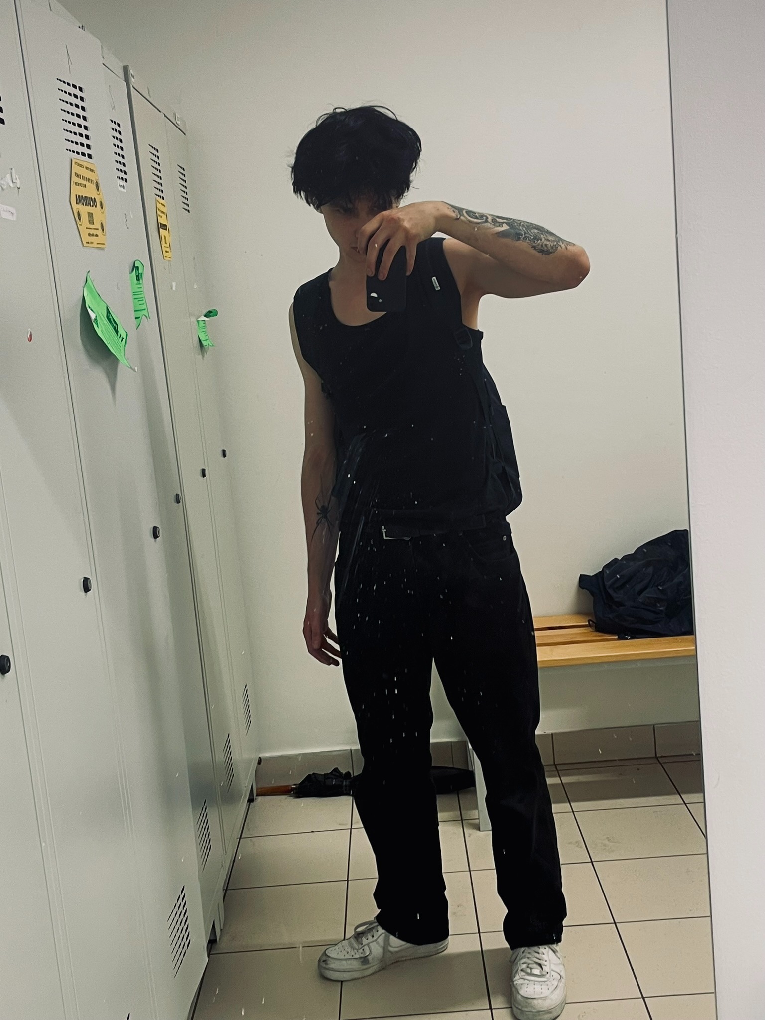 Mężczyzna w czarnym ubraniu robi selfie w lustrze na tle szatni z metalowymi szafkami i ławką. Widoczne tatuaże na ramionach i białe buty sportowe.
