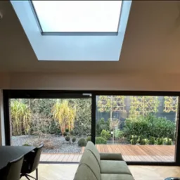 Jasne wnętrze z widokiem na zadbany ogród przez panoramiczne okno. Wnętrze doświetlone oknem dachowym. Widoczny fragment sofy i stołu z krzesłami.