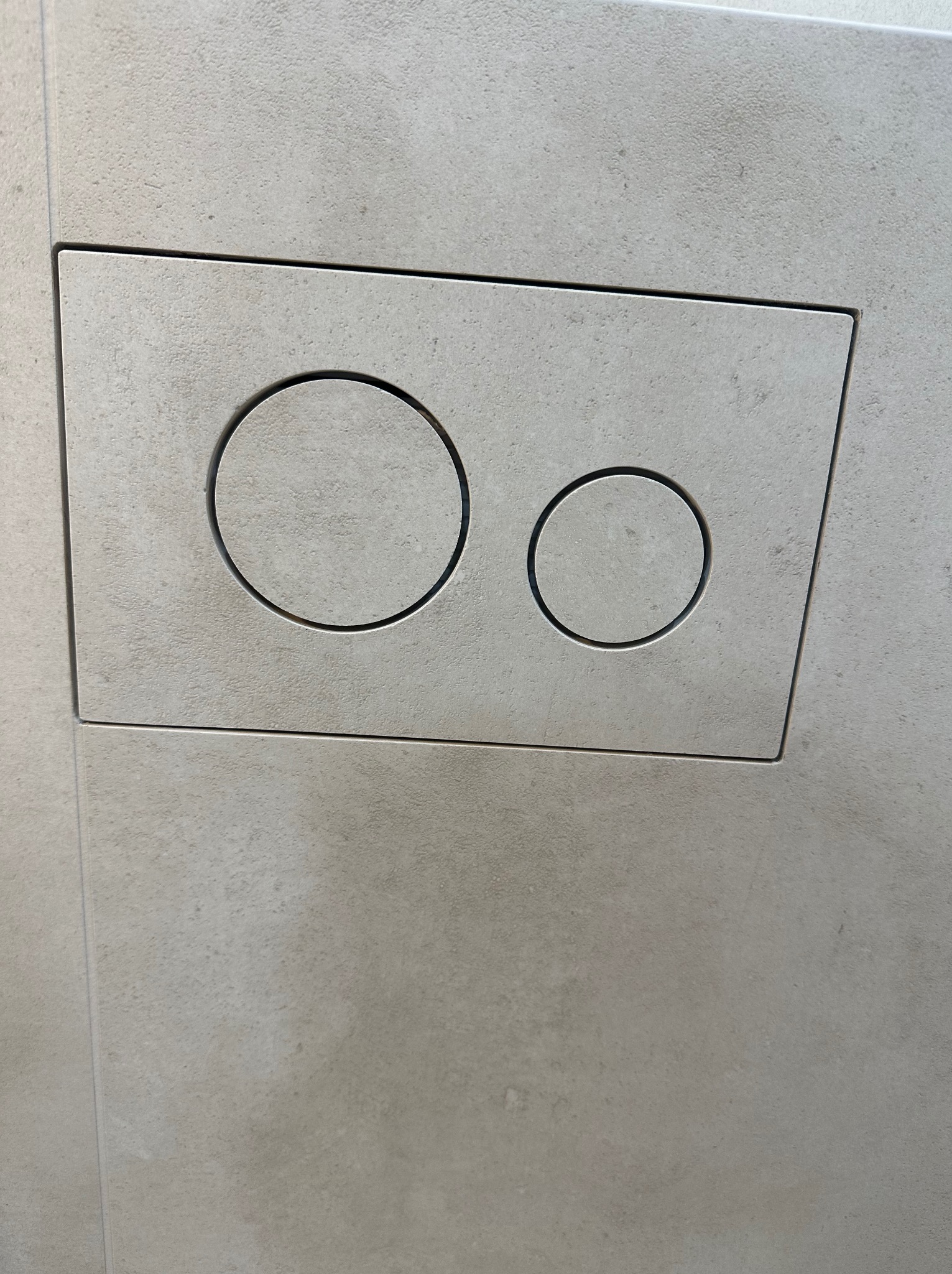 Przycisk spłuczki podtynkowej w łazience wykończonej jasnymi, matowymi płytkami. Minimalistyczny design z dwoma okrągłymi przyciskami o różnych rozmiarach.