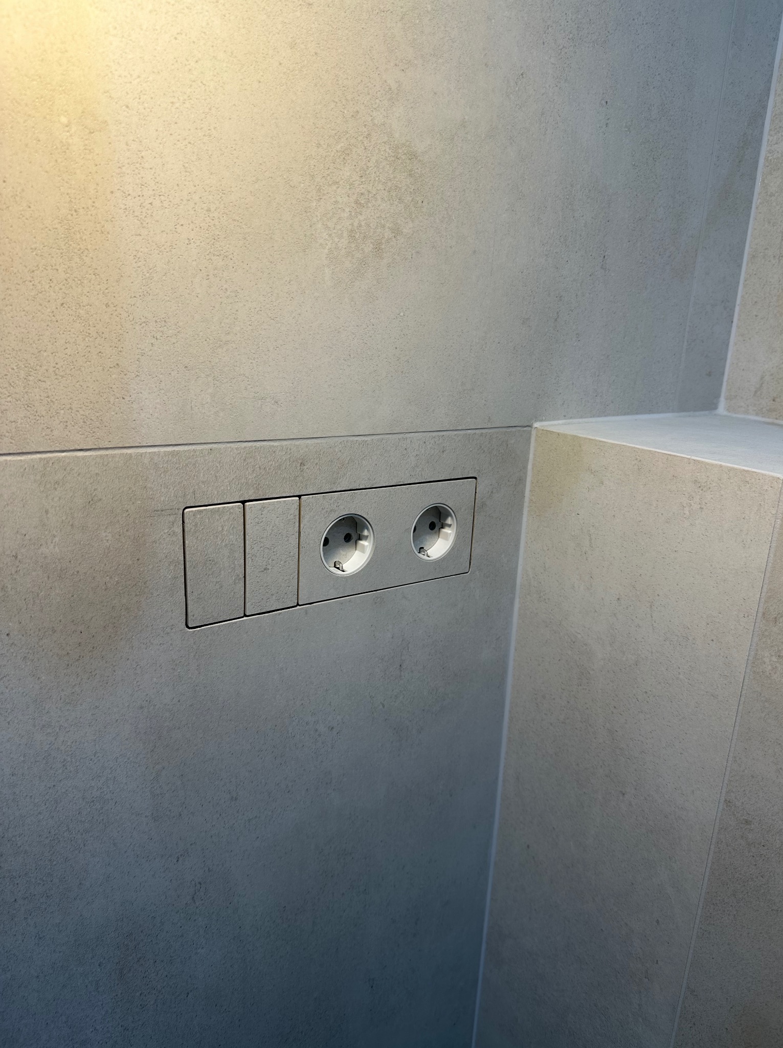 Szara, minimalistyczna łazienka z ukrytą spłuczką i gniazdkami elektrycznymi w płytkach. Nowoczesne wykończenie, stonowana kolorystyka, precyzyjne ułożenie płytek.